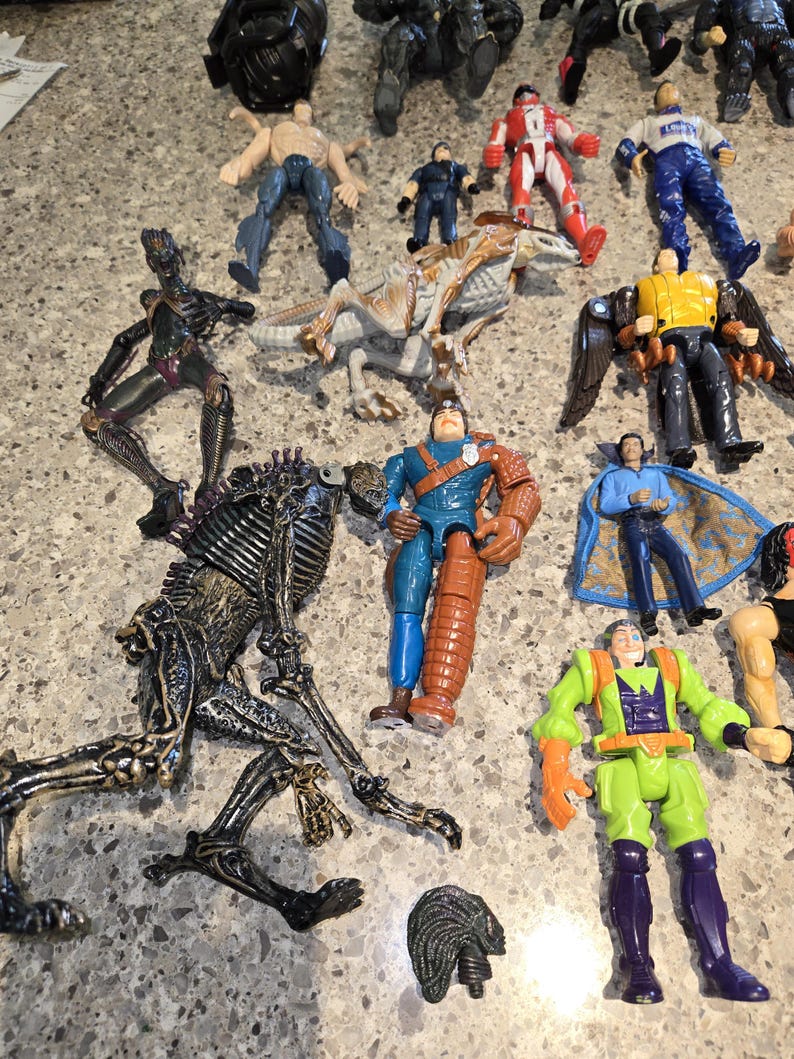 May include: A collection of various action figures, including a dinosaur, a robot, and several human-like figures. The figures are in various colors, including blue, red, green, and black. Some figures have wings or capes.