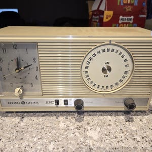 May include: Vintage General Electric radio-clock combo in a cream-colored case. Features a clock face on the left and a radio dial on the right, with AM/FM settings. Includes volume and tuning knobs. The clock shows the time.