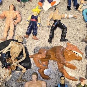 May include: A collection of action figures, including muscular humanoids, a skeleton figure, and a brown dinosaur-like creature. The figures are in various poses and feature a range of colors, including tan, blue, and black.