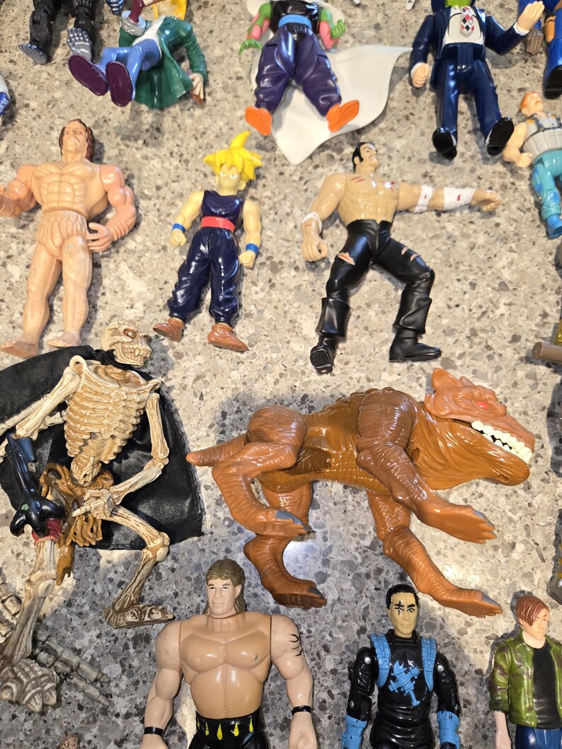 May include: A collection of action figures, including muscular humanoids, a skeleton figure, and a brown dinosaur-like creature. The figures are in various poses and feature a range of colors, including tan, blue, and black.