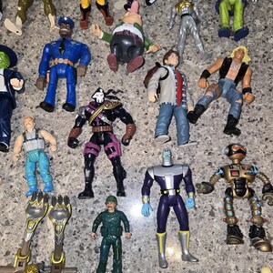 May include: A collection of vintage action figures, featuring various characters in vibrant colors. The figures include superheroes, robots, and other fantasy characters. Some figures are in shades of blue, green, yellow, and black.