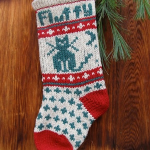 Könnte beinhalten: Ein weißer, roter und grüner gestrickter Weihnachtsstrumpf mit einem grünen Katzendesign. Der Strumpf hängt an einer Holzoberfläche.