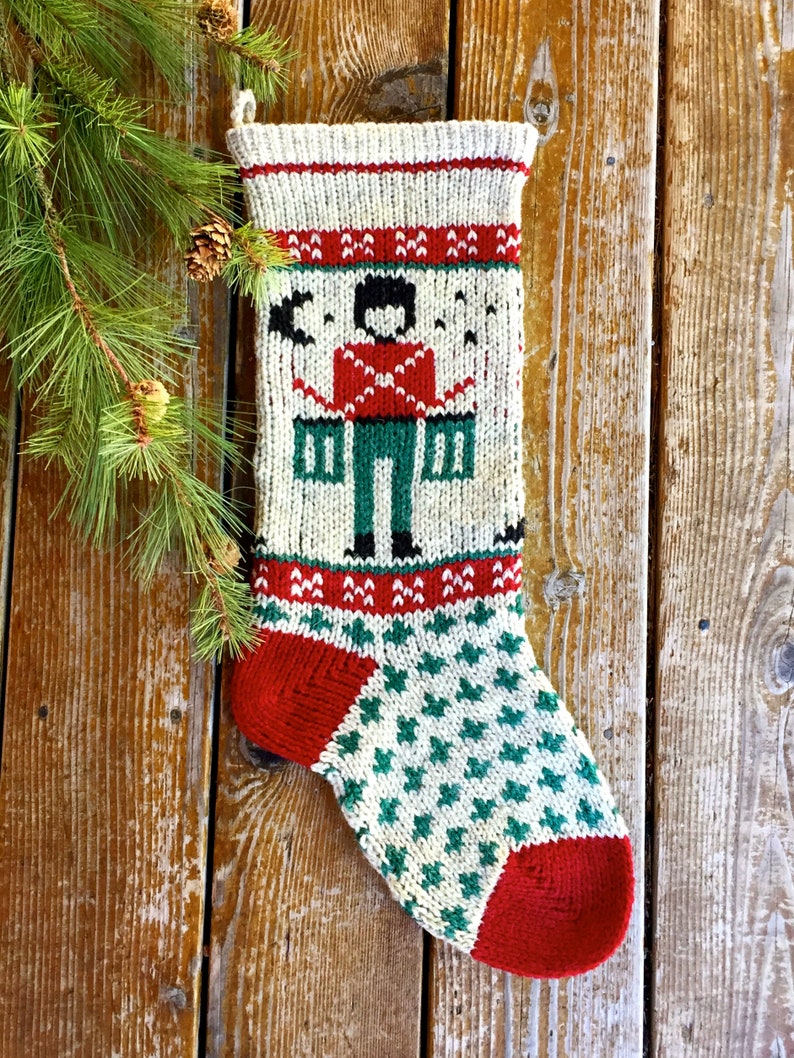 Peut inclure: Une chaussette de No&euml;l en tricot avec un motif rouge, blanc et vert. La chaussette pr&eacute;sente un motif de casse-noisette avec un chapeau noir et une veste rouge.
