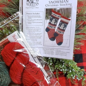 May include: A Christmas stocking knitting kit with red and green yarn, a pattern booklet, and a sprig of greenery. The booklet features a snowman design and the text "Annie's Woolens Snowman Christmas Stocking". The finished stocking size is 7" x 22".