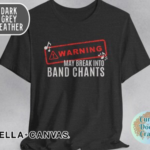 May include: Dark grey heather t-shirt with the text "WARNING MAY BREAK INTO BAND CHANTS" in white. The word "WARNING" is inside a red rectangle with musical notes. The shirt is from BELLA+CANVAS.