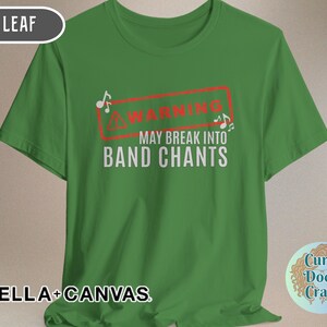 May include: Green t-shirt with the text "WARNING MAY BREAK INTO BAND CHANTS" in white, inside a red rectangle. The shirt also features musical notes. The brand name "BELLA + CANVAS" is printed at the bottom.
