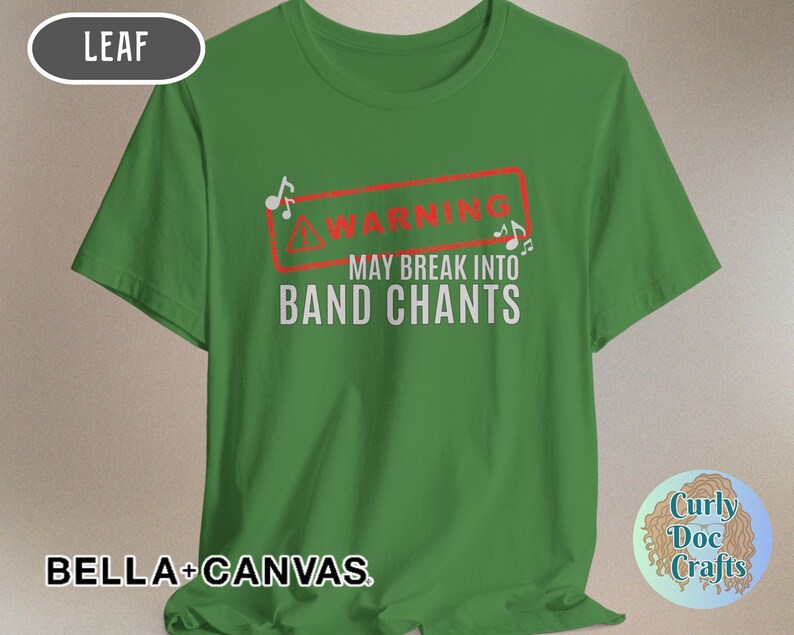 May include: Green t-shirt with the text "WARNING MAY BREAK INTO BAND CHANTS" in white, inside a red rectangle. The shirt also features musical notes. The brand name "BELLA + CANVAS" is printed at the bottom.