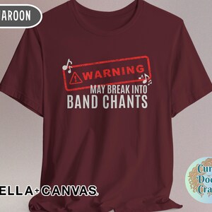 May include: Maroon t-shirt with the text "WARNING MAY BREAK INTO BAND CHANTS" in white. The word "WARNING" is inside a red rectangle with musical notes. The shirt is made of Bella+Canvas material.