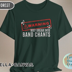 May include: Forest green t-shirt with the text "WARNING MAY BREAK INTO BAND CHANTS" in white. The word "WARNING" is inside a red rectangle with musical notes. The shirt is made of Bella+Canvas material.