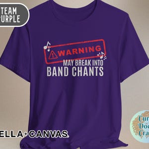 May include: A purple t-shirt with the text "WARNING MAY BREAK INTO BAND CHANTS" in white. The word "WARNING" is inside a red rectangle with musical notes. The shirt also has the words "TEAM PURPLE" and "BELLA + CANVAS".