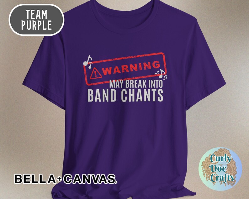 May include: A purple t-shirt with the text "WARNING MAY BREAK INTO BAND CHANTS" in white. The word "WARNING" is inside a red rectangle with musical notes. The shirt also has the words "TEAM PURPLE" and "BELLA + CANVAS".