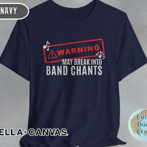 May include: Navy blue t-shirt with the text "WARNING MAY BREAK INTO BAND CHANTS" in white. The warning is in a red rectangle with musical notes. The shirt is made of Bella+Canvas material.