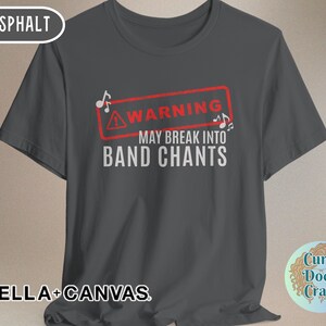 May include: Asphalt-colored t-shirt with the text "WARNING MAY BREAK INTO BAND CHANTS" in white. The warning is inside a red rectangle with musical notes. The shirt is from Bella+Canvas.