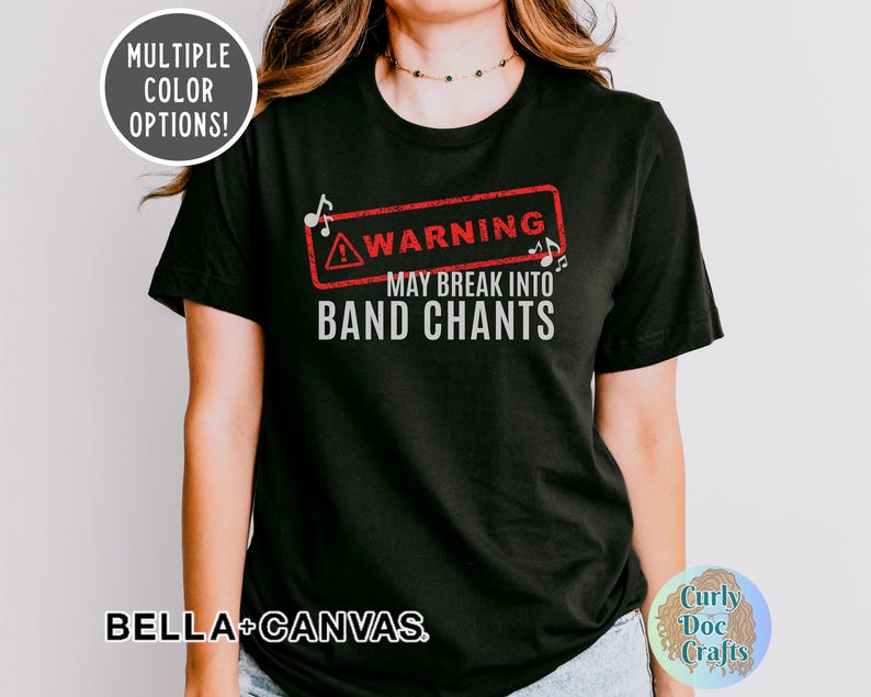 May include: Black t-shirt with the text "WARNING MAY BREAK INTO BAND CHANTS" in white and red. The shirt also features musical notes and a warning symbol. The brand name "BELLA+CANVAS" is printed at the bottom.
