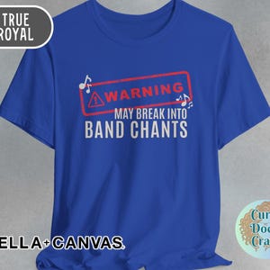 May include: Royal blue t-shirt with the text "WARNING MAY BREAK INTO BAND CHANTS" in white. The text is inside a red rectangle with musical notes. The shirt also has the text "BELLA + CANVAS".