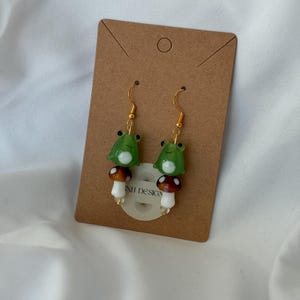 May include: A pair of handmade earrings featuring a green frog and a mushroom design. The frog has black eyes and sits atop a brown and white mushroom. The earrings hang from gold-colored hooks. The earrings are displayed on a brown card.
