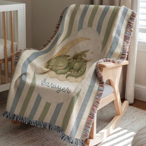 May include: A woven throw blanket featuring vertical stripes in muted blue, green, and cream. The center displays an oval graphic of a green dragon sleeping on a crescent moon and clouds, with the name Sawyer written in cursive script below.