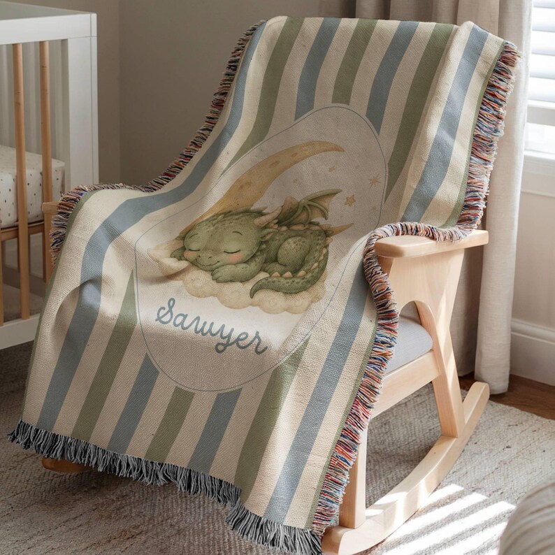 May include: A woven throw blanket featuring vertical stripes in muted blue, green, and cream. The center displays an oval graphic of a green dragon sleeping on a crescent moon and clouds, with the name Sawyer written in cursive script below.