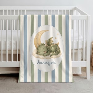 May include: A baby blanket featuring vertical stripes in cream, sage green, and dusty blue. The center displays an oval frame containing a sleeping green dragon resting on a cloud beneath a crescent moon and stars, with the name Sawyer printed below.