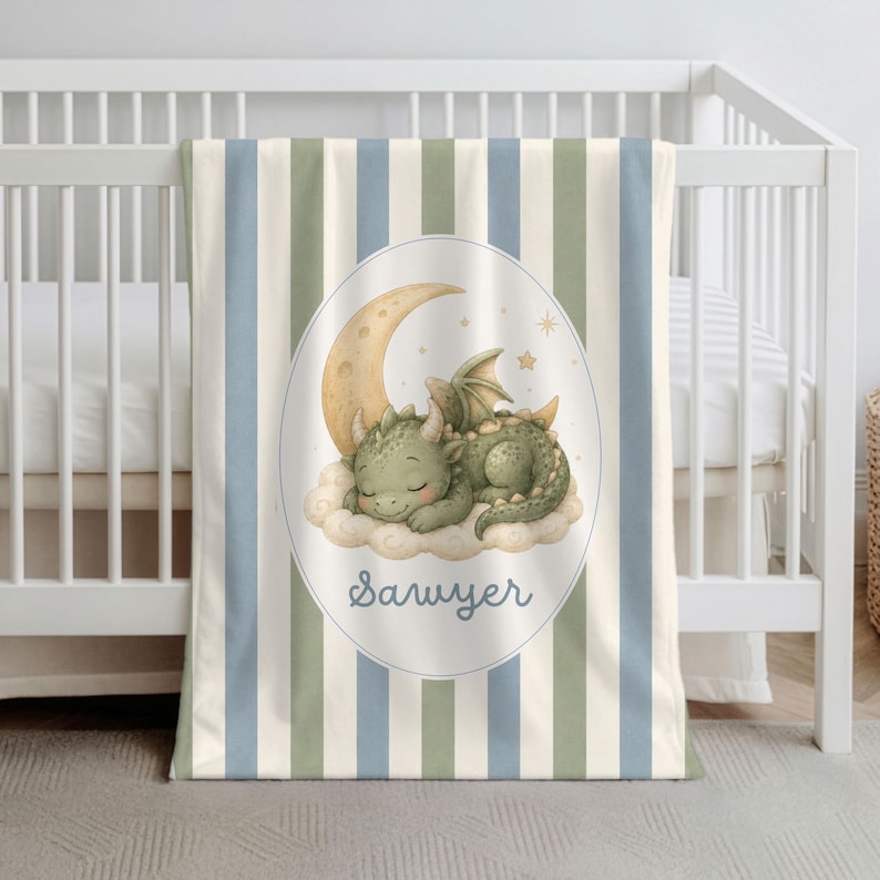 May include: A baby blanket featuring vertical stripes in cream, sage green, and dusty blue. The center displays an oval frame containing a sleeping green dragon resting on a cloud beneath a crescent moon and stars, with the name Sawyer printed below.