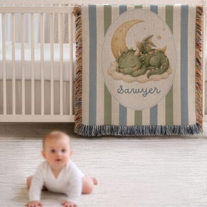 May include: A woven throw blanket featuring vertical blue and green stripes with a central oval graphic of a green dragon sleeping on a cloud beneath a crescent moon. The name Sawyer is printed in blue script below the dragon. The edges are fringed.