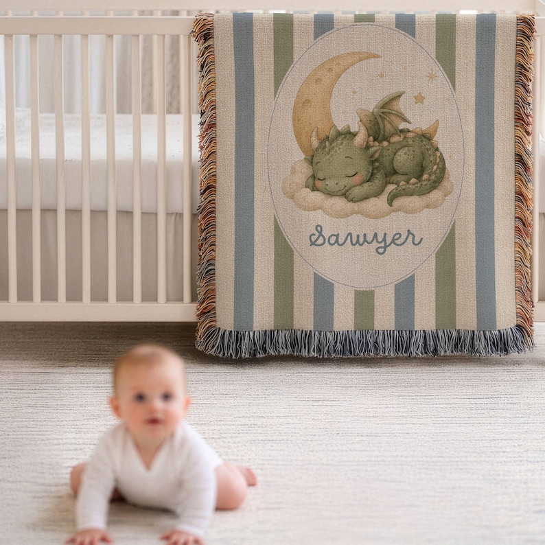 May include: A woven throw blanket featuring vertical blue and green stripes with a central oval graphic of a green dragon sleeping on a cloud beneath a crescent moon. The name Sawyer is printed in blue script below the dragon. The edges are fringed.