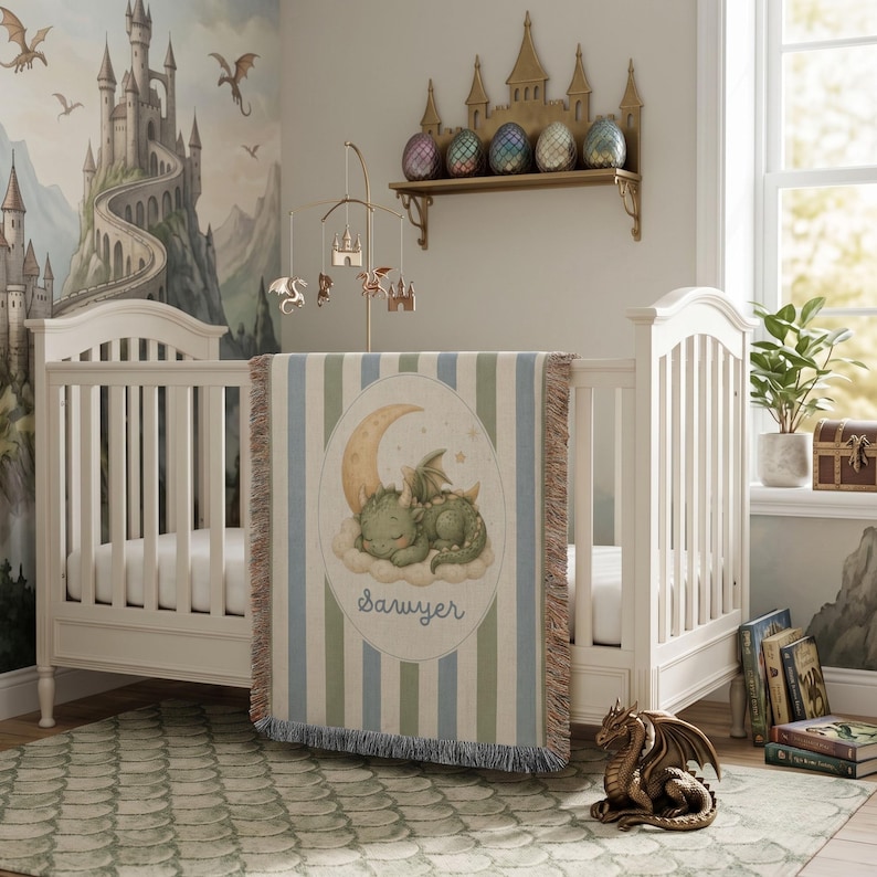 May include: A woven blanket featuring a central oval illustration of a green dragon sleeping on a crescent moon, with the name Sawyer printed below. The blanket has blue and white vertical stripes and a fringed edge, draped over a white crib.