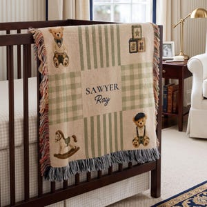 May include: A woven throw blanket featuring a beige and sage green plaid and striped pattern. The blanket displays the text "SAWYER RAY" in the center, surrounded by illustrations of two teddy bears, a rocking horse, and stacked toy blocks.