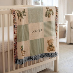 May include: A woven throw blanket featuring a grid pattern in shades of sage green and cream. The design includes illustrations of a vintage airplane, a toy car, a mallard duck, and a teddy bear, with the name SAWYER printed in the center.
