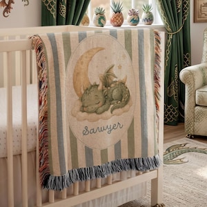 May include: A woven throw blanket featuring a green dragon sleeping on a cloud under a crescent moon. The blanket has vertical stripes in muted green, blue, and cream, with a fringed edge. The name Sawyer is printed in cursive below the dragon.