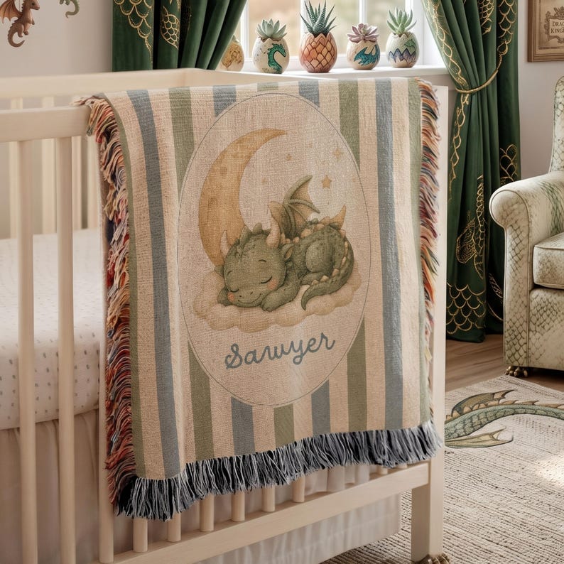 May include: A woven throw blanket featuring a green dragon sleeping on a cloud under a crescent moon. The blanket has vertical stripes in muted green, blue, and cream, with a fringed edge. The name Sawyer is printed in cursive below the dragon.