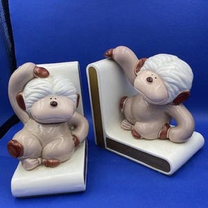 May include: A pair of ceramic bookends featuring a whimsical monkey design. The monkeys are brown and white with a fluffy texture and are holding books. The bookends are a great way to add a touch of fun to any bookshelf.