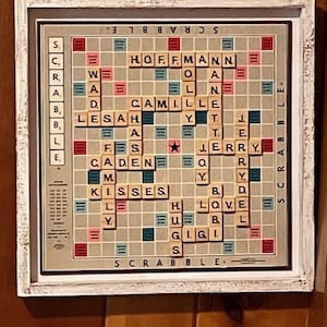 May include: A framed Scrabble board with a white distressed frame. The board features personalized words like "HOFFMANN", "CAMILLE", and "KISSES". The game board is in a neutral color palette with red, blue, and green accents.