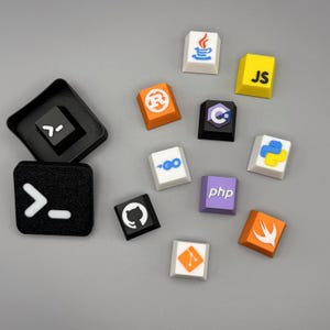 May include: A collection of colorful custom keycaps featuring various programming language logos, including Java, JavaScript, Python, and C++. The keycaps are in various colors, such as white, yellow, orange, and purple. Two black keycaps with white symbols are also present.