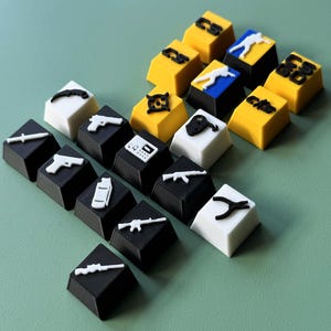 Może przedstawiać: Zestaw 16 niestandardowych klawiszy z r&oacute;żnymi symbolami i ikonami związanymi z grą Counter-Strike. Klawisze są czarne, białe i ż&oacute;łte z białymi ikonami. Klawisze są ułożone w kształcie piramidy.