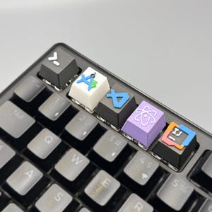 以下が含まれることがあります： カスタムキーキャップが付いた黒いメカニカルキーボードのクローズアップ。キーキャップには、緑色のアクセントが付いた白い「A」、青い「A」、紫色の原子記号、オレンジと青のアイコンなど、さまざまなカラフルなシンボルとアイコンが表示されています。キーボードは明るい灰色の表面にあります。