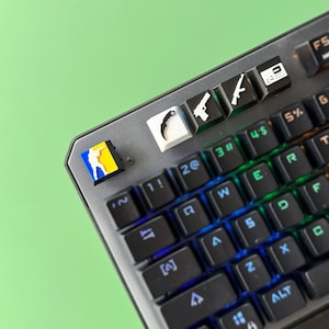 Może przedstawiać: Czarna mechaniczna klawiatura z podświetleniem RGB. Klawiatura ma klawisze z r&oacute;żnymi symbolami, w tym pistoletem, nożem i ikoną gracza. Klawiatura znajduje się na zielonej powierzchni.