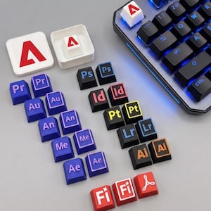 May include: A collection of custom mechanical keyboard keycaps featuring Adobe software icons. The set includes blue, black, and red keys with white, blue, pink, yellow, and orange lettering representing various creative applications and the Adobe logo.