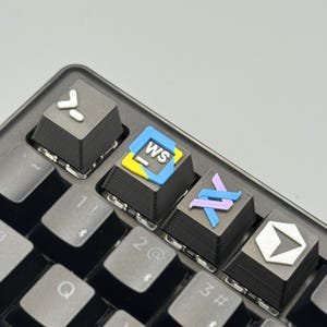 以下が含まれることがあります： 黒いメカニカルキーボードのクローズアップ。カスタムキーキャップが装着されており、白い矢印、「WS」の文字、抽象的な形など、カラフルなデザインとシンボルが特徴です。キーボードは灰色の表面にあります。