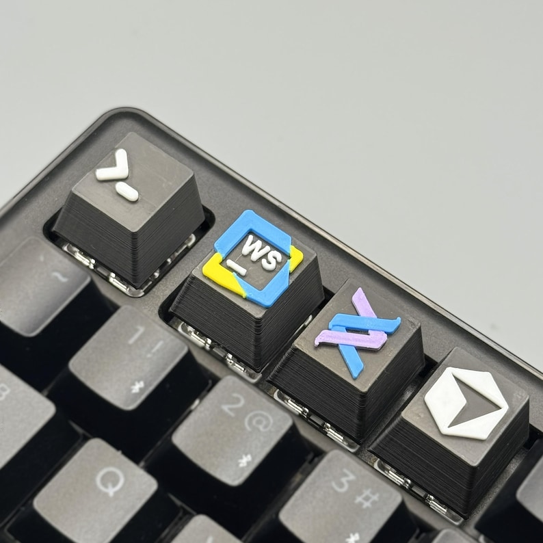 以下が含まれることがあります： 黒いメカニカルキーボードのクローズアップ。カスタムキーキャップが装着されており、白い矢印、「WS」の文字、抽象的な形など、カラフルなデザインとシンボルが特徴です。キーボードは灰色の表面にあります。