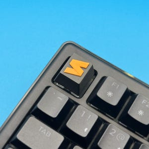 以下が含まれることがあります： オレンジ色の「M」キーキャップが付いた黒いメカニカルキーボードのクローズアップ。キーボードには、白い文字と記号が付いた灰色のキーがあります。背景は無地の青色です。