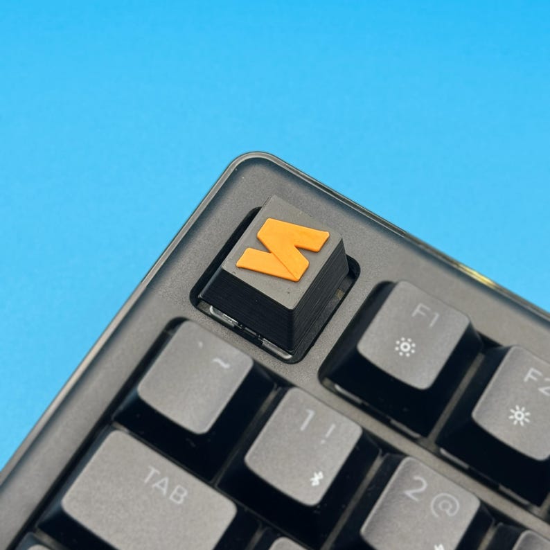 以下が含まれることがあります： オレンジ色の「M」キーキャップが付いた黒いメカニカルキーボードのクローズアップ。キーボードには、白い文字と記号が付いた灰色のキーがあります。背景は無地の青色です。