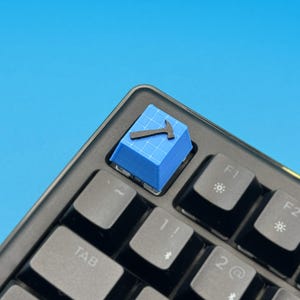 以下が含まれることがあります： 青いデザインと黒いハンマーアイコンが特徴のカスタムキーボードキーキャップ。キーキャップは黒いキーボードに取り付けられており、他の灰色のキーも見えます。背景は単色の青色です。