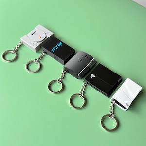 May include: A set of six keychains featuring different video game consoles. The keychains are made of black and white plastic and feature the logos of Sony PlayStation and Nintendo. The keychains are attached to silver key rings.