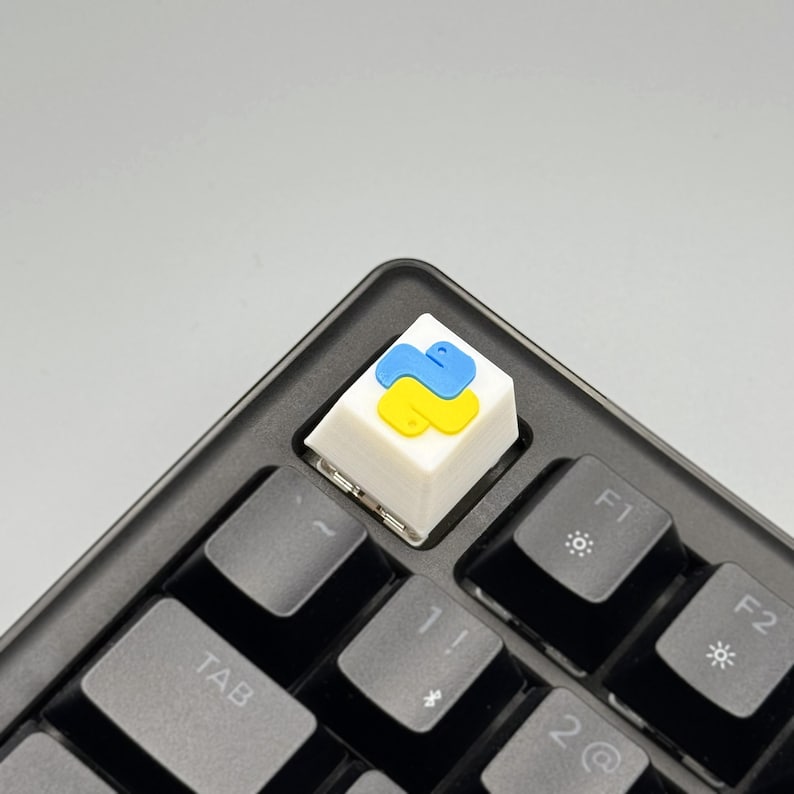 Puede incluir: Una tecla blanca con el logotipo de Python azul y amarillo instalada en un teclado negro. La tecla es cuadrada y se encuentra entre las teclas F1 y Tab. El logotipo de Python es una serpiente estilizada.