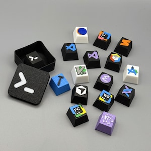 Könnte beinhalten: Eine Sammlung von individuellen Tastaturkappen in verschiedenen Farben, mit Symbolen und Icons. Die Tastenkappen sind schwarz, weiß, blau und lila, mit Designs wie einem Hammer, einem Kreis und einem stilisierten Atomsymbol. Ein schwarzer quadratischer Behälter enthält zusätzliche Tastenkappen.
