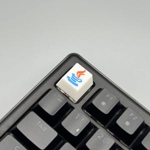 Puede incluir: Una tecla blanca con un gr&aacute;fico azul y naranja, instalada en un teclado negro. La tecla presenta un dise&ntilde;o estilizado, probablemente un logotipo o s&iacute;mbolo. Las teclas del teclado son negras y el fondo es gris claro.