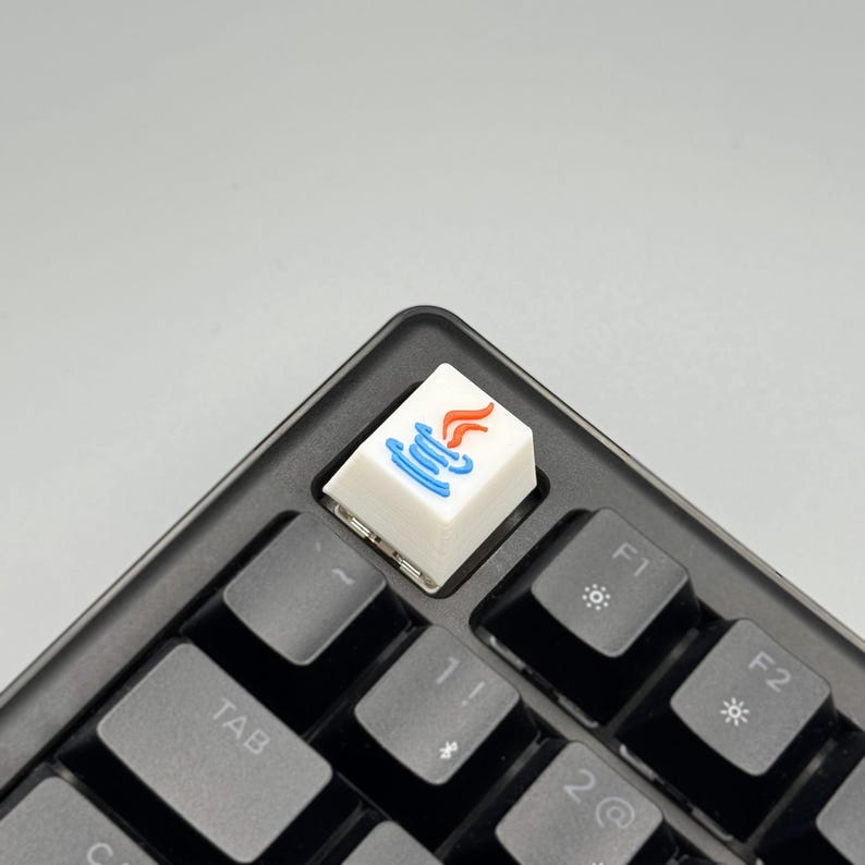 Puede incluir: Una tecla blanca con un gr&aacute;fico azul y naranja, instalada en un teclado negro. La tecla presenta un dise&ntilde;o estilizado, probablemente un logotipo o s&iacute;mbolo. Las teclas del teclado son negras y el fondo es gris claro.