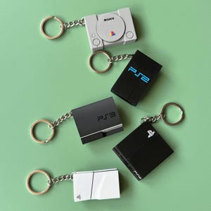 May include: Five different PlayStation console keychains. The keychains are all black and white and feature the PlayStation logo. The keychains are arranged in a row from left to right, with the oldest console on the left and the newest console on the right.