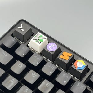 以下が含まれることがあります： カスタムキーキャップが付いた黒いメカニカルキーボードのクローズアップ。キーキャップには、緑と白のロゴが付いた白いキーキャップや、様式化された文字が付いた紫色のキーキャップなど、さまざまなカラフルなデザインとシンボルが表示されています。